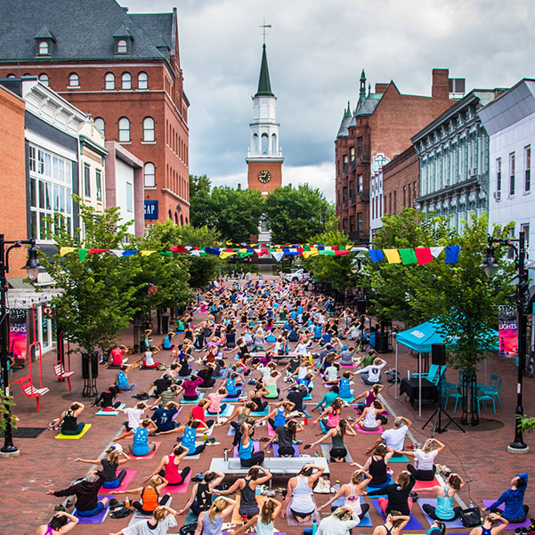 A beautiful aerial photo of Church street on a summer day. The street is hosting a community yoga event. A large group of women are sitting on yoga mats peforming a pose in unison, with the church tower in the background. 