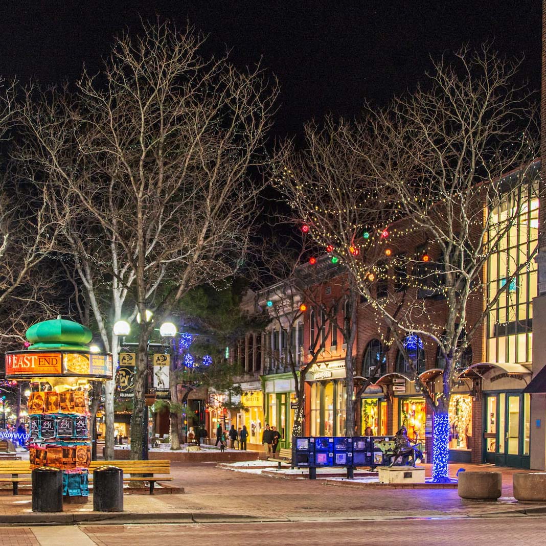 A winter night on Pearl street. Bare trees decorated with Christmas lights line the middle of the street. A bulletin board and newspaper stand are in the foreground, with few people walking and socializing by the storefronts in the distance.