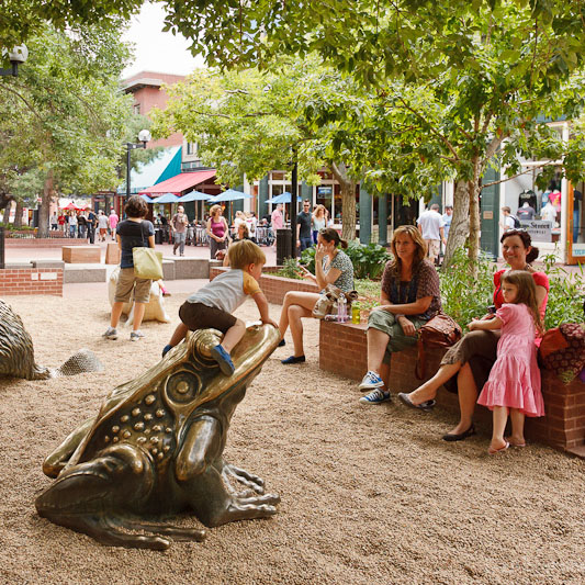 Children playing in Pearl street's sandbox. A boy is climbing a brass statue of a frog in the foreground, with a girl and mothers sitting on the side.