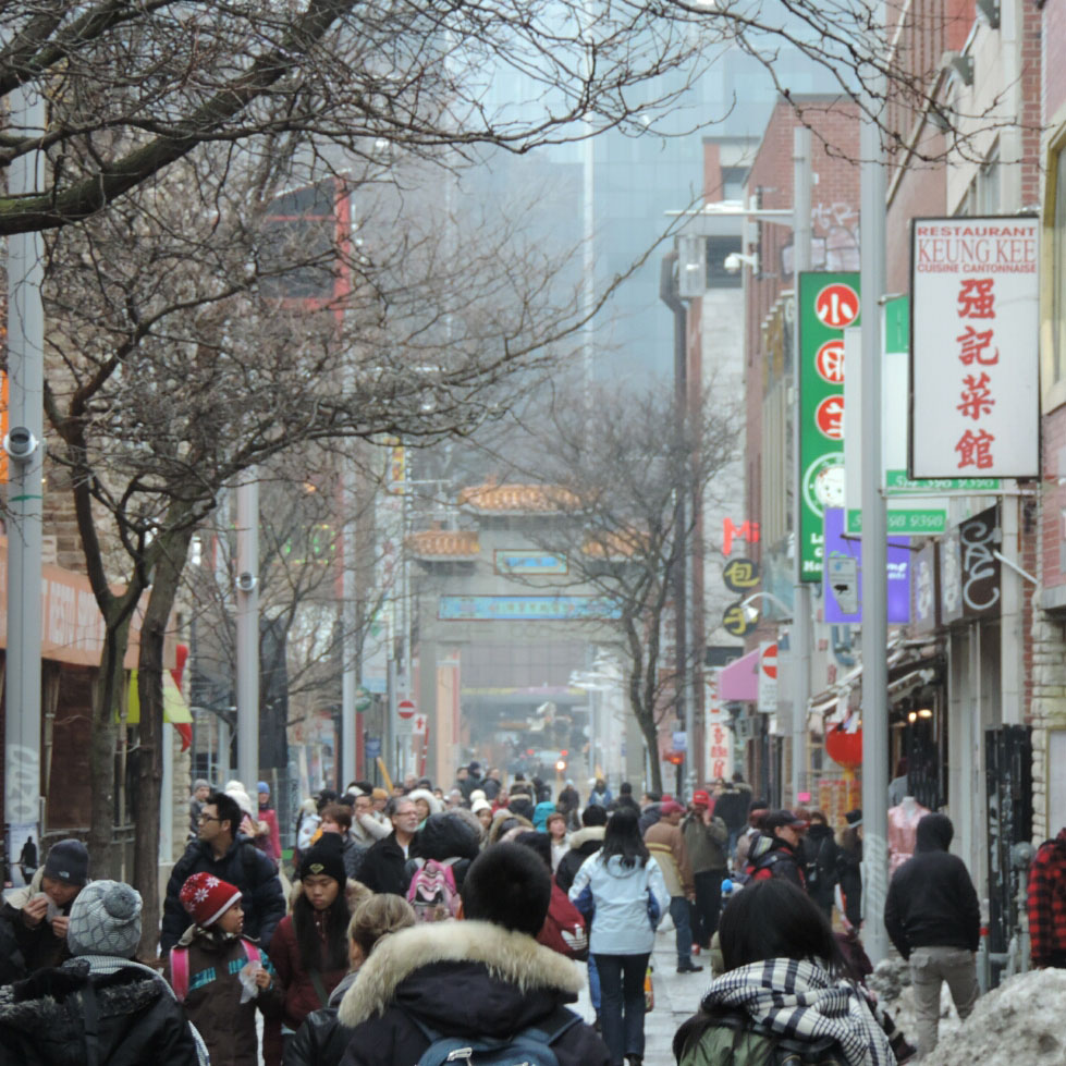 The busy pedestrian zone of de la Gauchetiere street in Chinatown on a hazy winter day. The gates to the neighbourhood and buildings downtown are seen in the distance. Restaurants and shops line the street with people eating and window shopping.