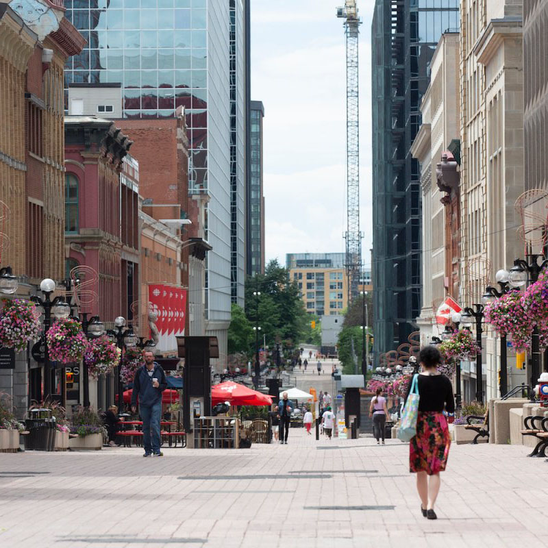 A woman walking on Sparks street. A variety of historic and modern buildings line the street. The ornate street lamps are decorated with bundles of pink flowers. Several other people are walking in the background, including a man holding a coffee.