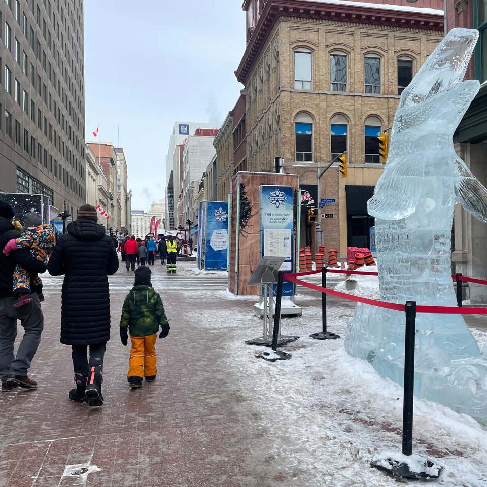A couple walking with their children on a winter day. To their side is a large ice sculpture of a fish rising from the water.