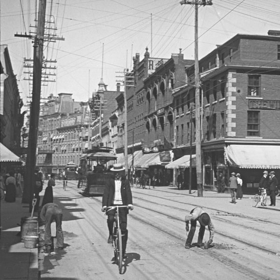 A black and white historic photo of Sparks Street. The street is lined with telephone poles and wires overhead. A man rides a bicycle in the foreground. A streetcar approches in the background with workers clearing the dirt road and its tracks of debris.
