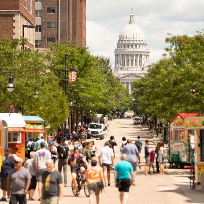 A tree-lined State street on a summer day. People are walking in different directions. In the background, a couple of delivery vehicles are parked to the side, with the state capitol building in the distance.