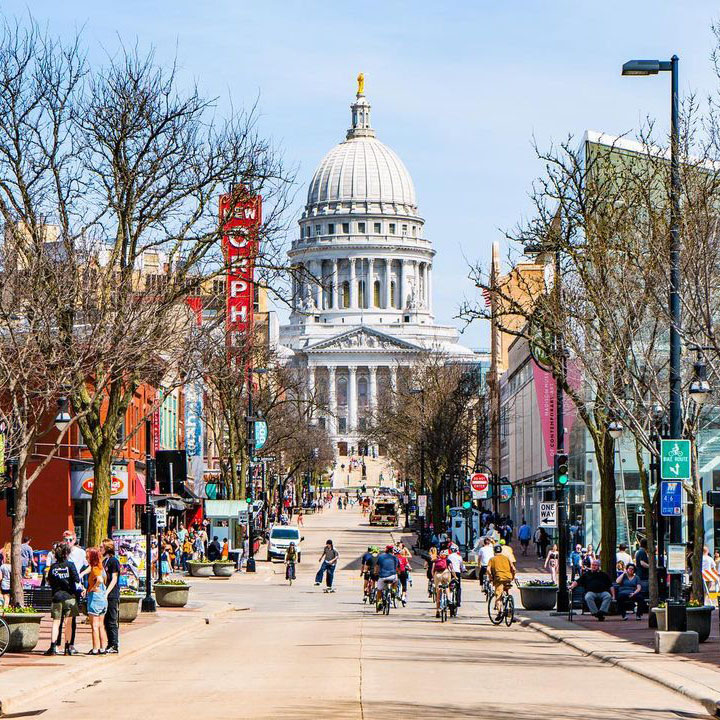 A view of the capitol building and its steps from State street. A group of cyclists and a skateboarder use the middle of the street, with many other people walking, sitting, and socializing on the raised sidewalks. 