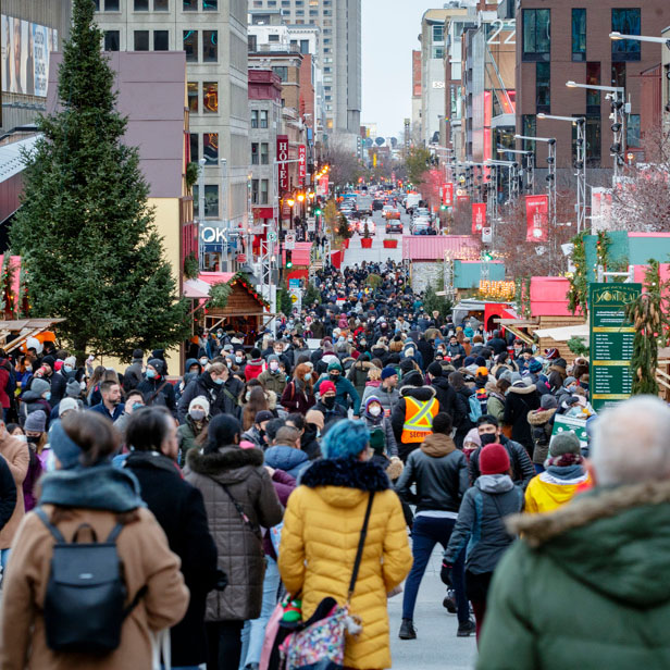 A crowded Sainte Catherine street open to pedestrians on a cold day. A number of food stalls, a pine tree and festive decorations are set up in the street. Car traffic is visible in the distance at the limits of the pedestrian zone.