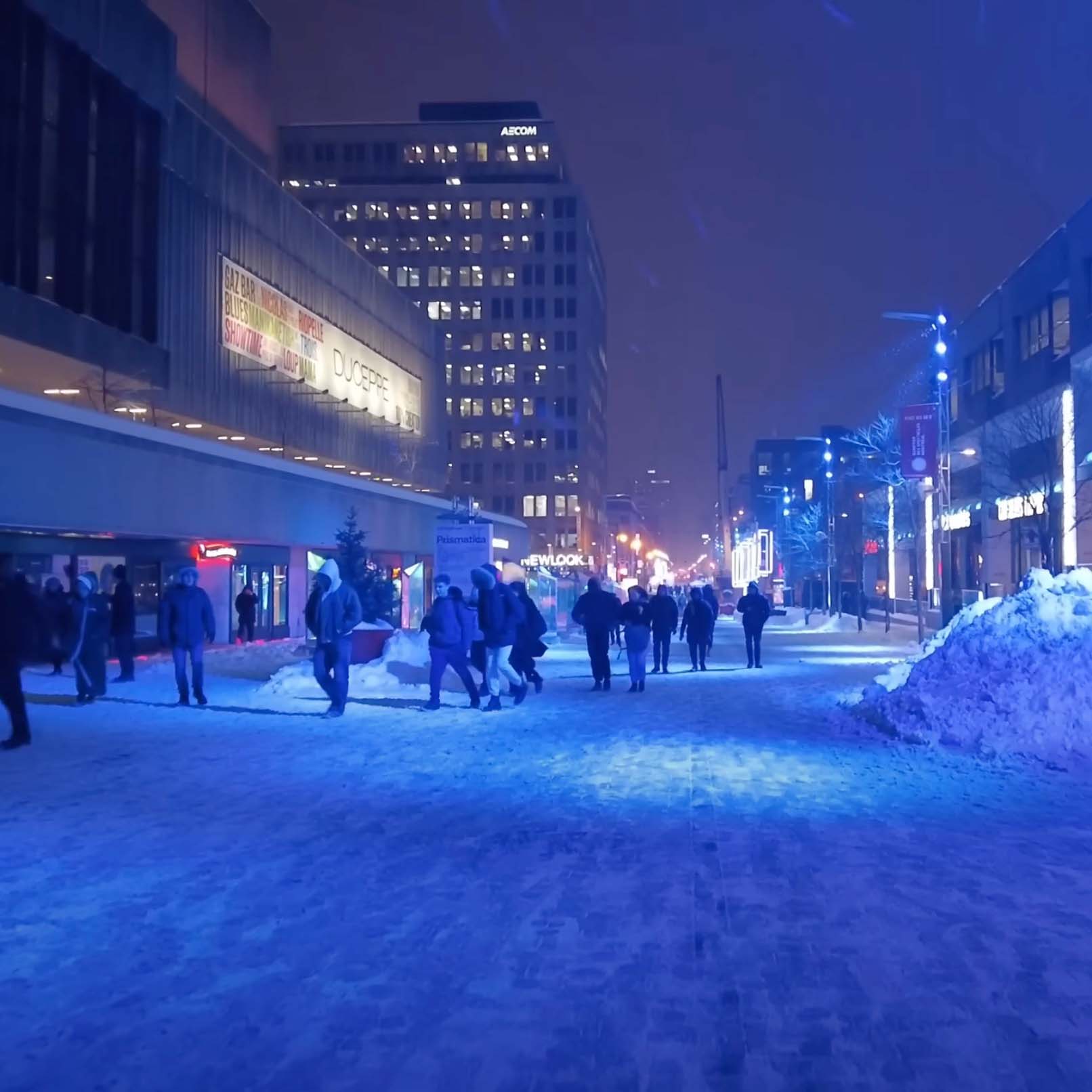 A winter night on the pedestrian zone of Saint Catherine street. The street is illuminated with intense blue lights. People walk in front of the entrance of Place des arts. A mound of plowed snow is pushed neatly to the side.