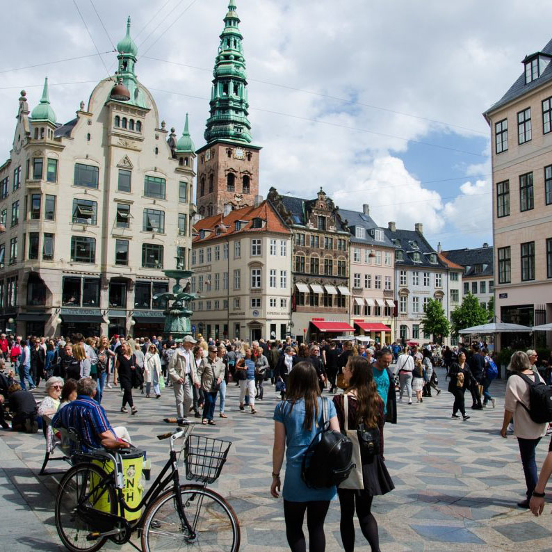 A busy, crowded day on the central square of StrÃ¸get. A bicycle is parked in the forground with buildings and a church tower in the background.